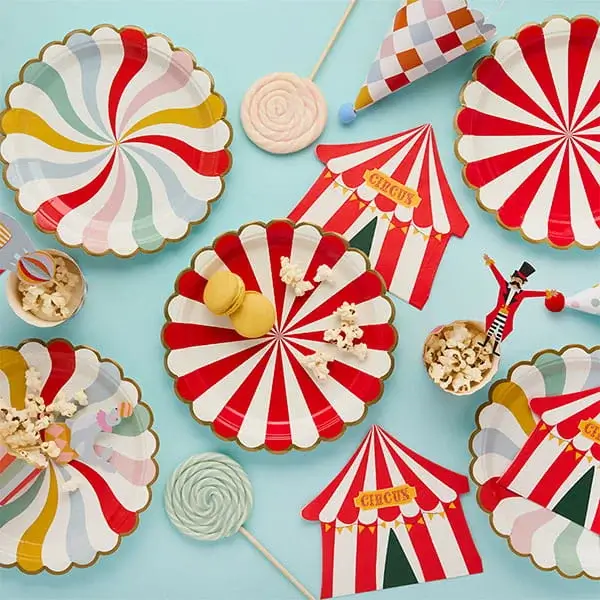 Circus-themed party tableware with red and white striped plates, tent decorations, popcorn, and colorful sweets on a blue background